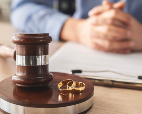 Wooden judge’s gavel and two gold wedding rings on a desk with a blurred client in the background, symbolizing high-end divorce and complex asset division in Montgomery County Pennsylvania.
