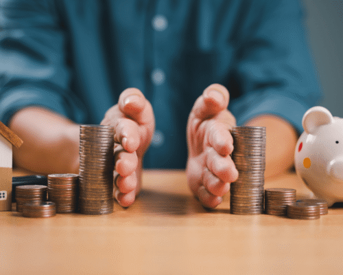 Close-up of hands dividing stacks of coins between a miniature house and a piggy bank, symbolizing tax law divorce asset division PA. The image highlights capital gains divorce 2024 PA, tax on retirement transfers PA, and asset liquidation tax rules divorce under updated Pennsylvania law.