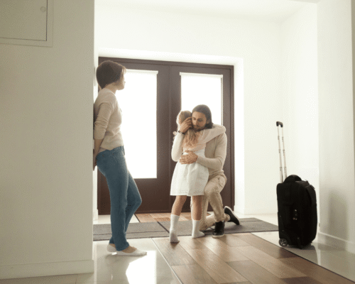 Father kneels to hug his daughter at the doorway while the mother stands nearby and a suitcase rests by the door, symbolizing child relocation custody Montgomery County PA. The image conveys parent relocation Pennsylvania law, custody relocation request PA, parenting plans long distance, and move-away custody cases.
